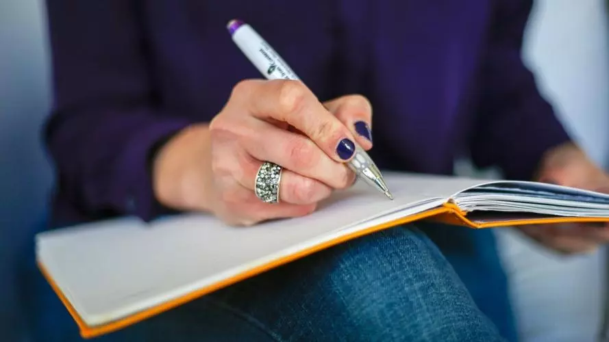 Person wearing dark nail polish writing in a notebook that is balanced on their knee.