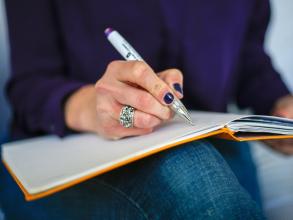 Person wearing dark nail polish writing in a notebook that is balanced on their knee.